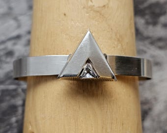 May include: A silver ring with a geometric triangular design. The ring features a polished band and a prominent triangle shape at the center, with a small, clear gemstone set within the triangle. The ring is displayed on a wooden ring holder.