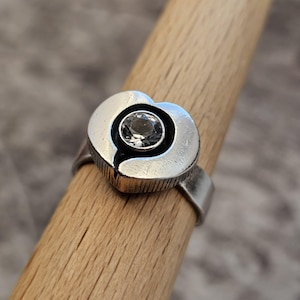 May include: A silver-toned ring with a heart-shaped design. The ring features a central, clear gemstone set within the heart. The band has a textured finish, and the ring is displayed on a wooden dowel.