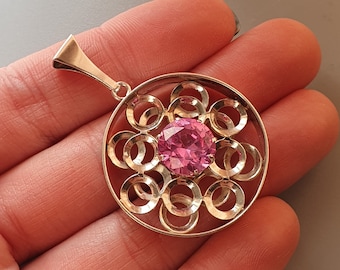 May include: A silver pendant with a pink gemstone surrounded by a circular design of interlocking rings.