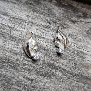 May include: A pair of silver-toned earrings with a leaf-like design. Each earring features a curved, textured shape with a small, round, clear gemstone at the base. The earrings are displayed on a weathered, gray wooden surface.