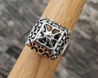 May include: A silver-colored ring with a square, openwork design. The ring features a geometric pattern on the sides and a central, star-like shape within the square. The ring is displayed on a wooden dowel.