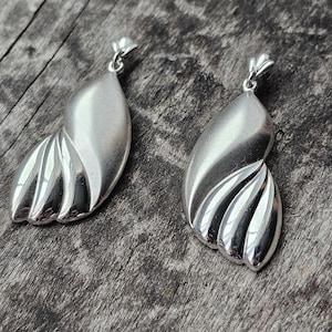 May include: A pair of silver-toned metal pendants featuring a smooth, teardrop-shaped upper section and a lower portion defined by three distinct, polished, curved ridges. Each pendant includes a small metal bail at the top for attachment.