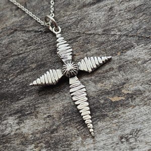 May include: A silver-toned metal cross pendant suspended from a matching thin chain. The cross features four elongated arms with a textured, ribbed surface pattern and a small, circular, sunburst-style centerpiece where the arms intersect.