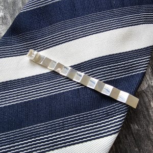 May include: A silver tie clip with a rectangular design. The tie clip is lying on a blue and white striped fabric.