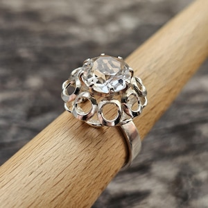 May include: A silver ring featuring a large, clear, faceted gemstone. The gemstone is surrounded by a decorative setting with open circular designs. The ring is displayed on a wooden dowel against a blurred background.