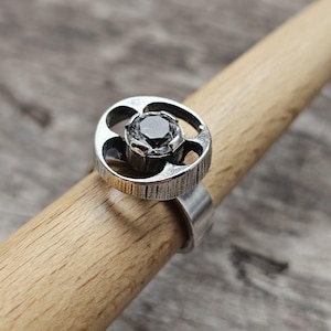 May include: A silver ring with a large, dark gray gemstone set in a circular, openwork design. The ring has a textured band and is displayed on a wooden dowel. The ring's design is modern and geometric.