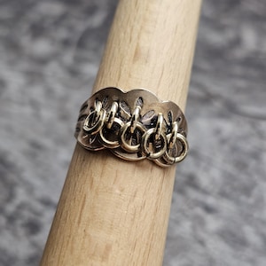 May include: A bronze-colored ring with a decorative, scalloped band and multiple dangling ring accents. The ring is displayed on a wooden dowel against a blurred gray background. The ring's design features intricate cut-out patterns.