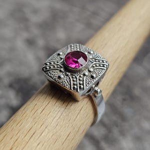 Op de afbeelding: Een zilveren ring met een vierkante voorkant met een sierlijk ontwerp en een ronde, magenta edelsteen. De ring wordt getoond op een houten deuvel, waarbij de ingewikkelde details en het vakmanschap worden benadrukt.