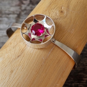Op de afbeelding: Zilveren armband met een cirkelvormig middelpunt met een levendige, ronde, magenta edelsteen. Het middelpunt heeft een open ontwerp met een stervormig patroon rond de edelsteen. De armband wordt getoond op een houten deuvel.