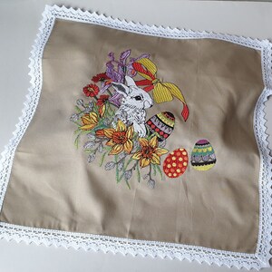 May include: A beige fabric square with a white lace border features an embroidered design of a white rabbit surrounded by colorful flowers and Easter eggs.