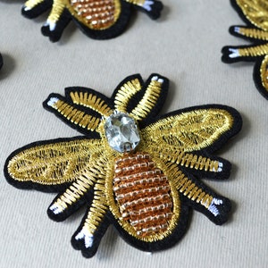 May include: Gold embroidered bee patch with a clear rhinestone and brown beaded body. The bee is on a black felt background.