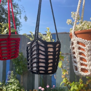 Op de afbeelding: Drie hangende plantenpotten gemaakt van gehäkeld macramé. De potten zijn rood, zwart en wit. Elke pot heeft een plant die erin groeit.