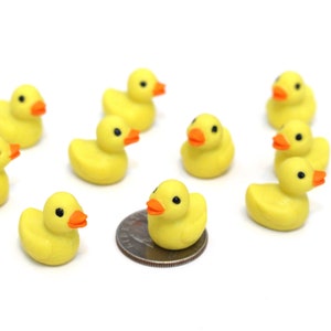 May include: A group of nine yellow rubber duck toys with orange beaks and black eyes. One duck is sitting on a silver US quarter.