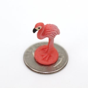 May include: A pink flamingo figurine standing on a silver coin. The flamingo is facing left and has a black beak and black eyes.