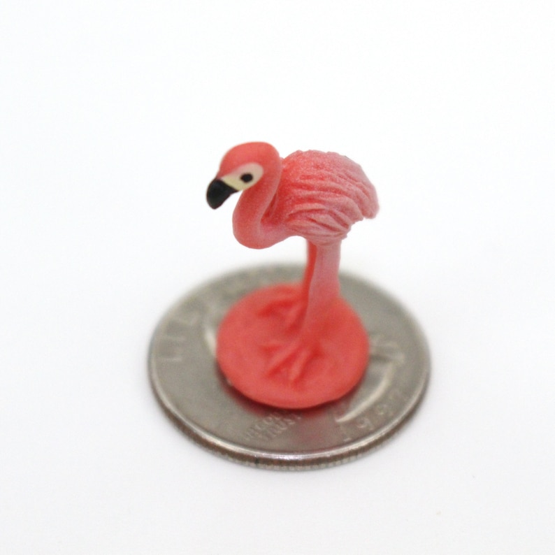 May include: A pink flamingo figurine standing on a silver coin. The flamingo is facing left and has a black beak and black eyes.