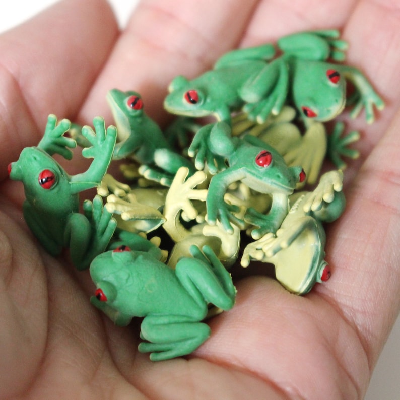 May include: A group of small green plastic frog figurines with red eyes. The frogs are in a variety of poses, some with their legs outstretched and others with their legs tucked in. The frogs are all about 2 inches long.