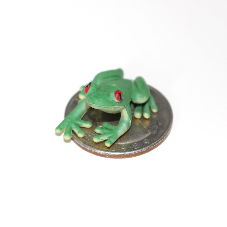 May include: A small green frog figurine with red eyes sitting on a US quarter.