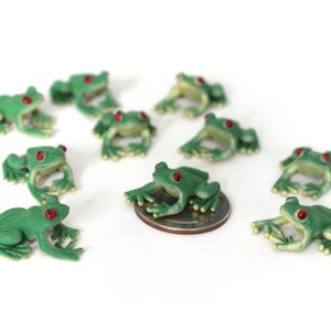 May include: Nine green plastic frog figurines with red eyes. The frogs are posed in different positions and are all about 1 inch in size. One frog is sitting on a US quarter.