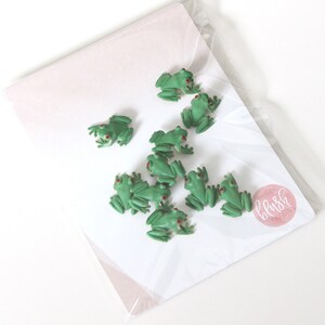 May include: A set of ten green frog figurines with red eyes. The frogs are arranged on a white background with a pink circle logo that says "blush studio".