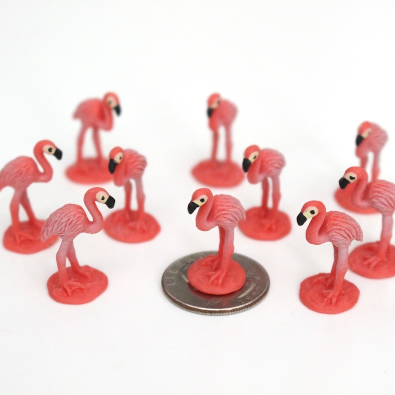 May include: A group of nine pink plastic flamingo figurines, each standing on a small pink base. The figurines are arranged in a circle around a silver US quarter.