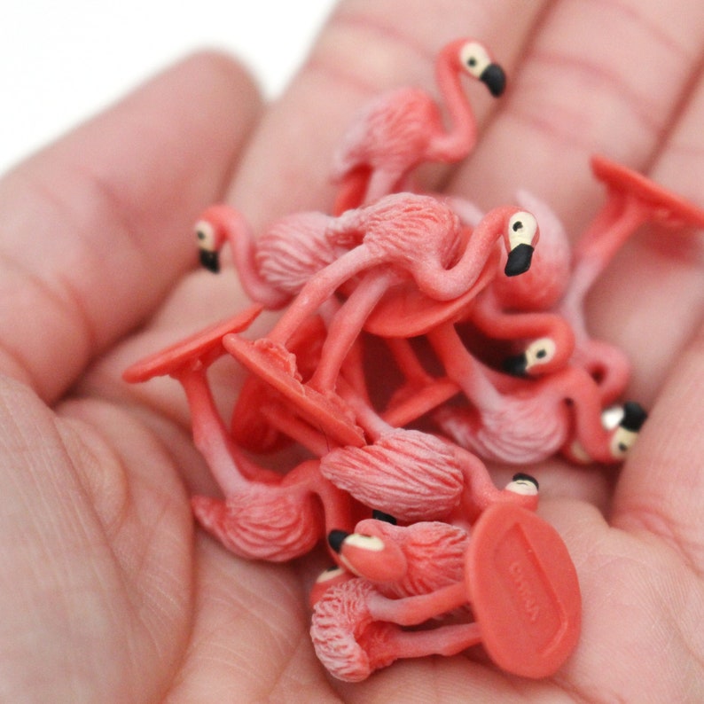 May include: A group of pink plastic flamingo figurines, each with a black beak and a white base. The figurines are arranged in a pile, with some of them standing upright and others lying on their sides.