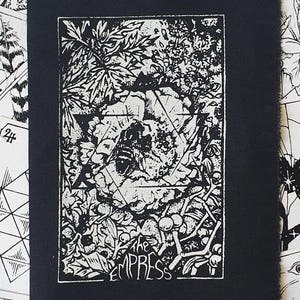 May include: A black and white print of "The Empress" tarot card. The image shows a detailed illustration of a natural scene with leaves, a geometric shape, and the card's title. The print is set against a backdrop of other tarot cards.