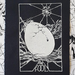 May include: A black and white tarot card print featuring an egg cracking open, with the words "THE FOOL" at the bottom. The print is surrounded by other tarot cards with various illustrations.