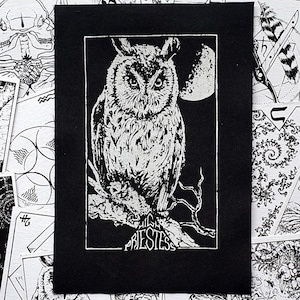 May include: A black and white linocut print featuring an owl perched on a branch, with a full moon behind it. The print is set in a black mount, with the word "PRIESTESS" at the bottom. Other black and white illustrations are in the background.