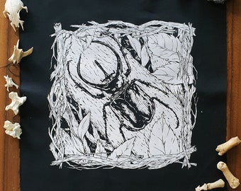 Könnte beinhalten: Schwarzer Stoff-Patch mit einem weißen Design eines Käfers in einem Blatt-Rahmen. Der Käfer hat große Hörner und ist von Blättern und Ranken umgeben.