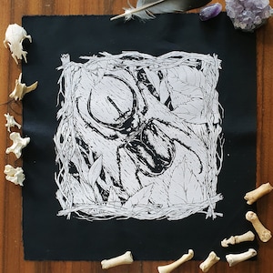 Könnte beinhalten: Schwarzer Stoff-Patch mit einem weißen Design eines Käfers in einem Blatt-Rahmen. Der Käfer hat große Hörner und ist von Blättern und Ranken umgeben.