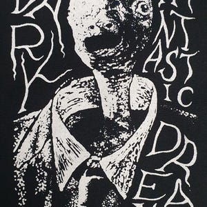 Könnte beinhalten: Schwarzes Grafik-T-Shirt mit einer weißen Illustration eines grotesken Gesichts und den Worten "DARK REALMS" und "FANTASTIC DREAMS" in stilisierten Buchstaben. Das Design zeigt ein Hemd mit Kragen.