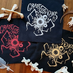 Może przedstawiać: Trzy czarne łatki z tkaniny z białym, czerwonym i złotym wzorem stworzenia z wieloma oczami z nadrukiem "Grant Us Eyes" na każdej łatce.