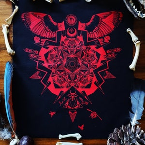 May include: Red and black geometric print on black fabric with a winged symbol at the top and a mandala-like design in the center. The print is surrounded by bones, feathers, and a pine cone.