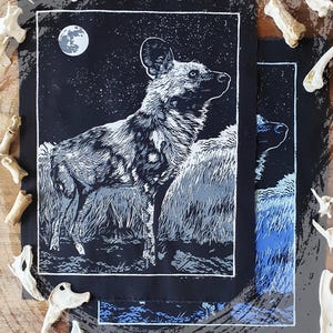 Könnte beinhalten: Schwarz-Weiß-Illustration eines Afrikanischen Wildhundes unter einem Sternenhimmel mit Vollmond. Das Kunstwerk befindet sich auf schwarzem Hintergrund mit weißem Rand. Ein weiteres Kunstwerk mit blauen und grauen Farbtönen ist ebenfalls zu sehen.