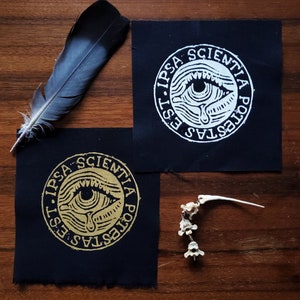 Op de afbeelding: Twee zwarte stoffen patches met de tekst "EST. IPSA SCIENTIA POTESTAS" rondom een oogontwerp. Eén patch heeft een gouden ontwerp, de andere wit. Een veer en een kleine botstructuur zijn ook aanwezig.