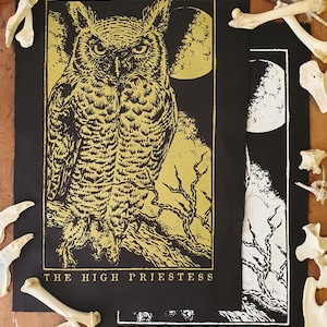 Könnte beinhalten: Zwei schwarze Stoffbahnen mit goldenen und weißen Illustrationen. Eine zeigt eine Eule mit Vollmond, die andere eine Mondlichtszene. Der Text "THE HIGH PRIESTESS" befindet sich am unteren Rand jedes Panels. Knochenfragmente und eine Feder umgeben die Panels.