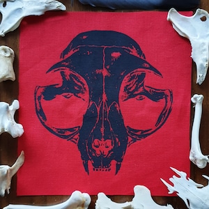 May include: A black and white printed fabric patch with a skull design. The skull is of a cat with large eye sockets and a prominent jaw. The patch is on a red background.