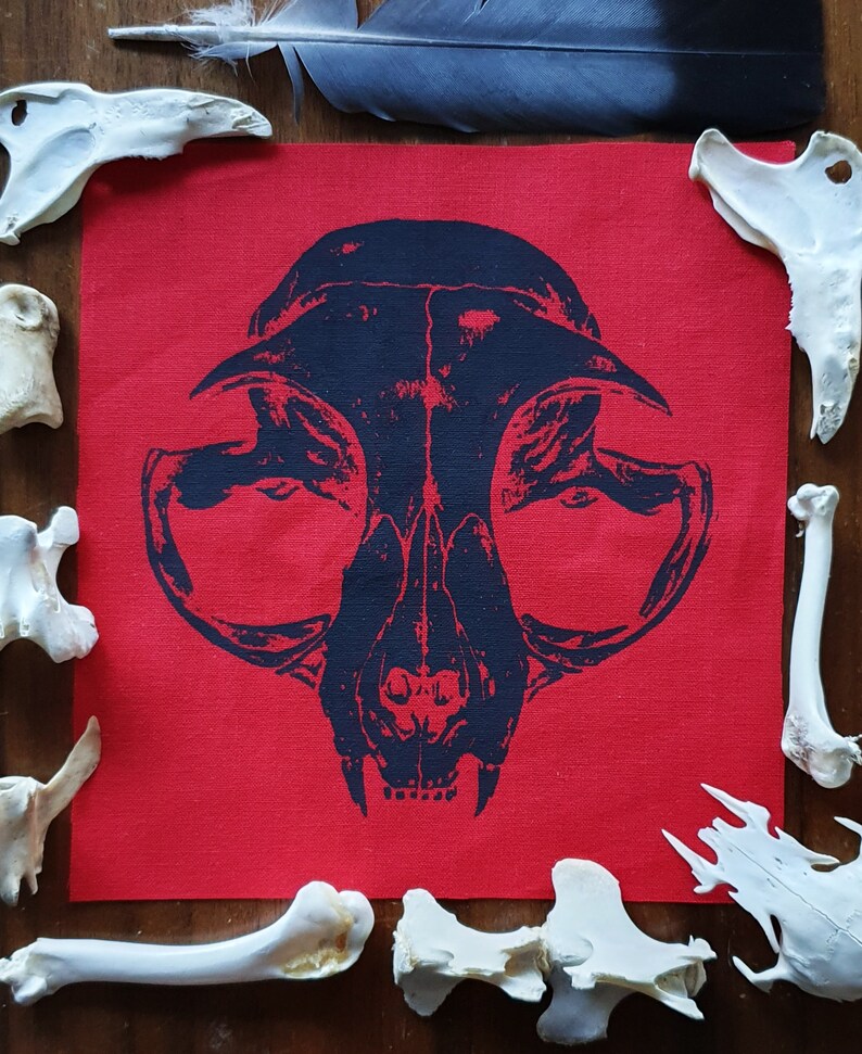 May include: A black and white printed fabric patch with a skull design. The skull is of a cat with large eye sockets and a prominent jaw. The patch is on a red background.