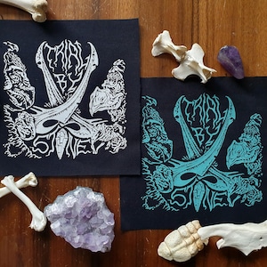 以下が含まれることがあります： 白い文字とターコイズブルーの文字で「Trial By Stone」と書かれた黒い布のパッチが2つ。パッチには、後ろに2本の剣が交差した頭蓋骨が描かれている。