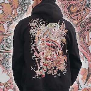 Op de afbeelding: Zwarte hoodie met een gedetailleerde, kleurrijke afbeelding op de rug. Het kunstwerk toont een complex ontwerp met rode, gouden en witte elementen, dat lijkt op een wezen of een abstracte vorm. De hoodie heeft een capuchon.