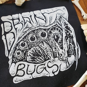 Op de afbeelding: Zwart vierkantige patch met witte grafische kunst en tekst. Het ontwerp toont een gedetailleerde, gestileerde illustratie van een wezen met meerdere ogen en organische vormen. De tekst "BEAST BUGS" is aan de onderkant gebogen.