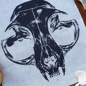 May include: Black and white printed image of an animal skull on a light gray fabric background. The skull is a close-up of the front of the skull, showing the teeth and eye sockets.