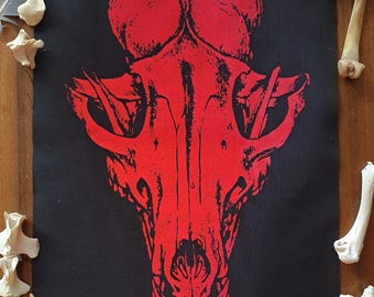 May include: A rectangular black fabric patch featuring a high-contrast, bright red screen-printed graphic of a canine skull. The skull design is centered on the fabric, displaying detailed bone structures, eye sockets, and nasal cavities.