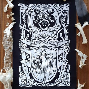 Peut inclure: Une estampe linogravure en noir et blanc d'un scarabée avec des détails complexes. Le scarabée est entouré d'une bordure décorative.