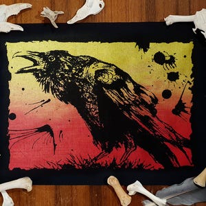 May include: A black raven illustration with an open beak, set against a gradient background of yellow and red. The artwork is framed in black and surrounded by animal bones. The raven's feathers are detailed, and the image has a textured, artistic feel.