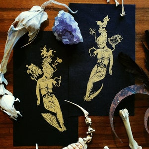 Op de afbeelding: Twee zwarte kunstprints met gouden illustraties van mensachtige figuren, samen met dierenbotten, veren en een paarse kristalcluster. De figuren hebben gestileerde kenmerken en vloeiende elementen. De compositie is gerangschikt op een houten oppervlak.