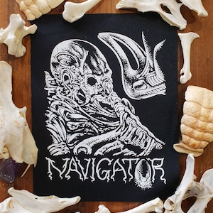 Pode incluir: Etiqueta bordada em preto e branco com uma ilustração detalhada de uma criatura alienígena esquelética com a palavra "NAVIGATOR" impressa abaixo.
