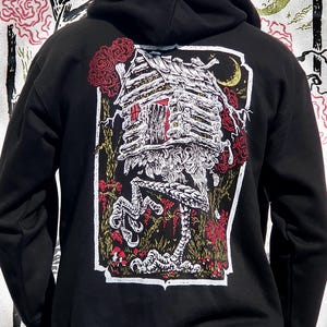 Puede incluir: Sudadera negra con cremallera y capucha con un estampado gráfico de una casa con patas y una luna creciente en el fondo. La casa está hecha de troncos y tiene un techo rojo. El gráfico está rodeado por un marco blanco.
