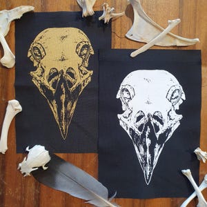 Könnte beinhalten: Zwei schwarze Stoffpatches mit einem weißen Vogelschädel-Design auf dem einen und einem goldenen Vogelschädel-Design auf dem anderen.
