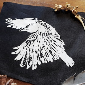 May include: A white silhouette of a bird with its wings spread out against a black fabric background.